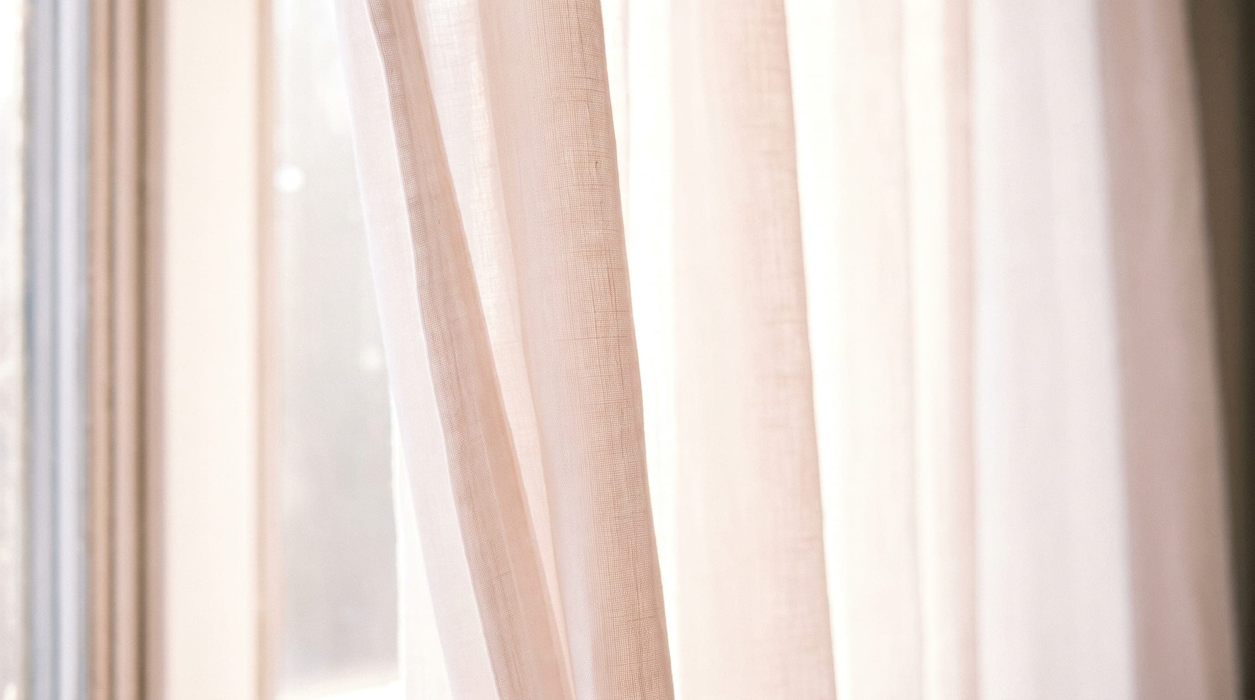 Sheer blush linen curtain glowing where daylight passes through a tall window.