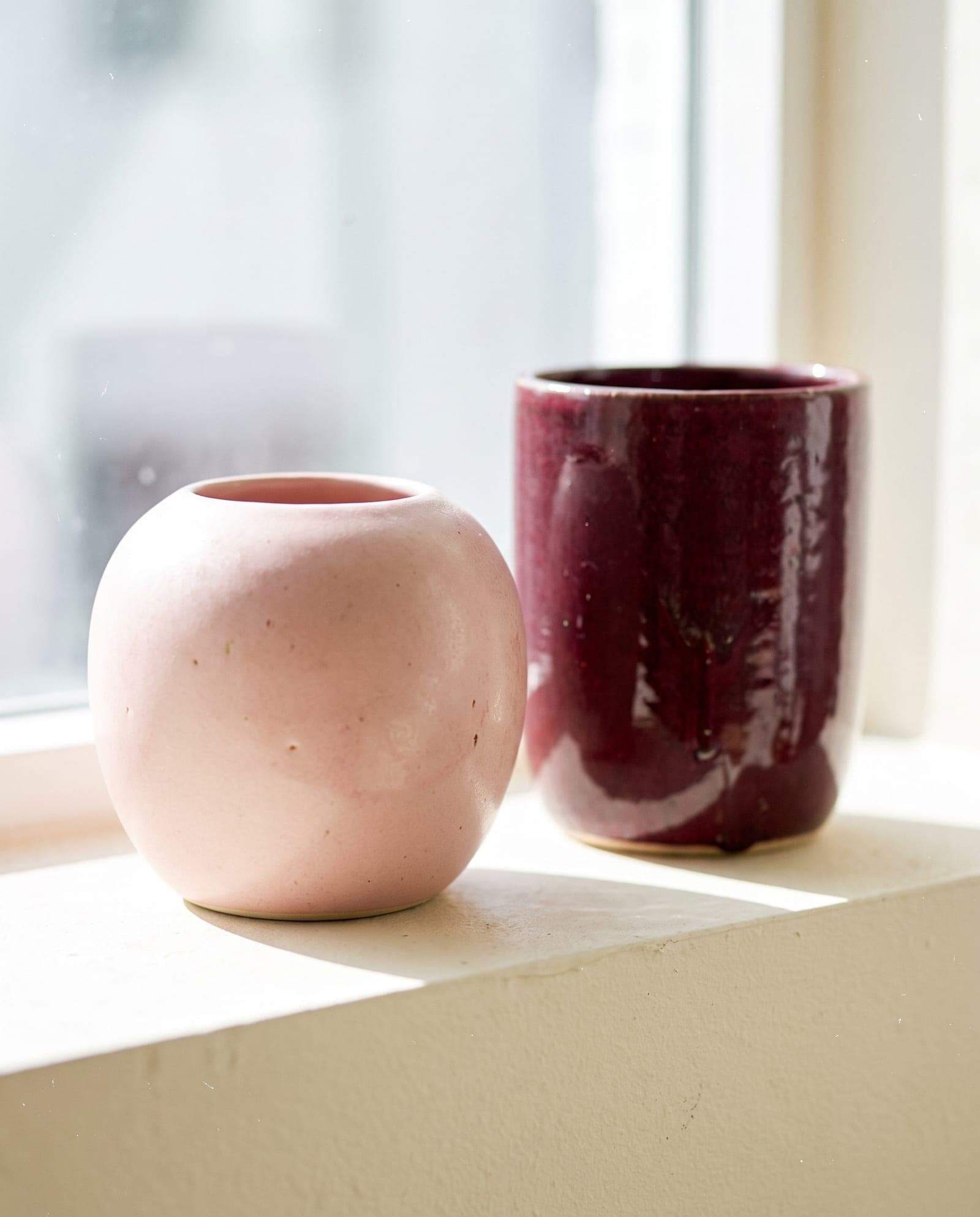 Two ceramic vessels in a blush glaze and a deep claret glaze, standing in clean daylight.