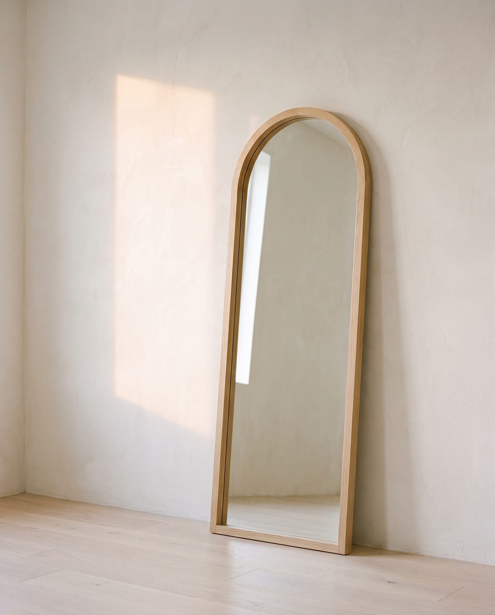 A tall arched oak floor mirror against a warm plaster wall, reflecting only the empty sunlit room.
