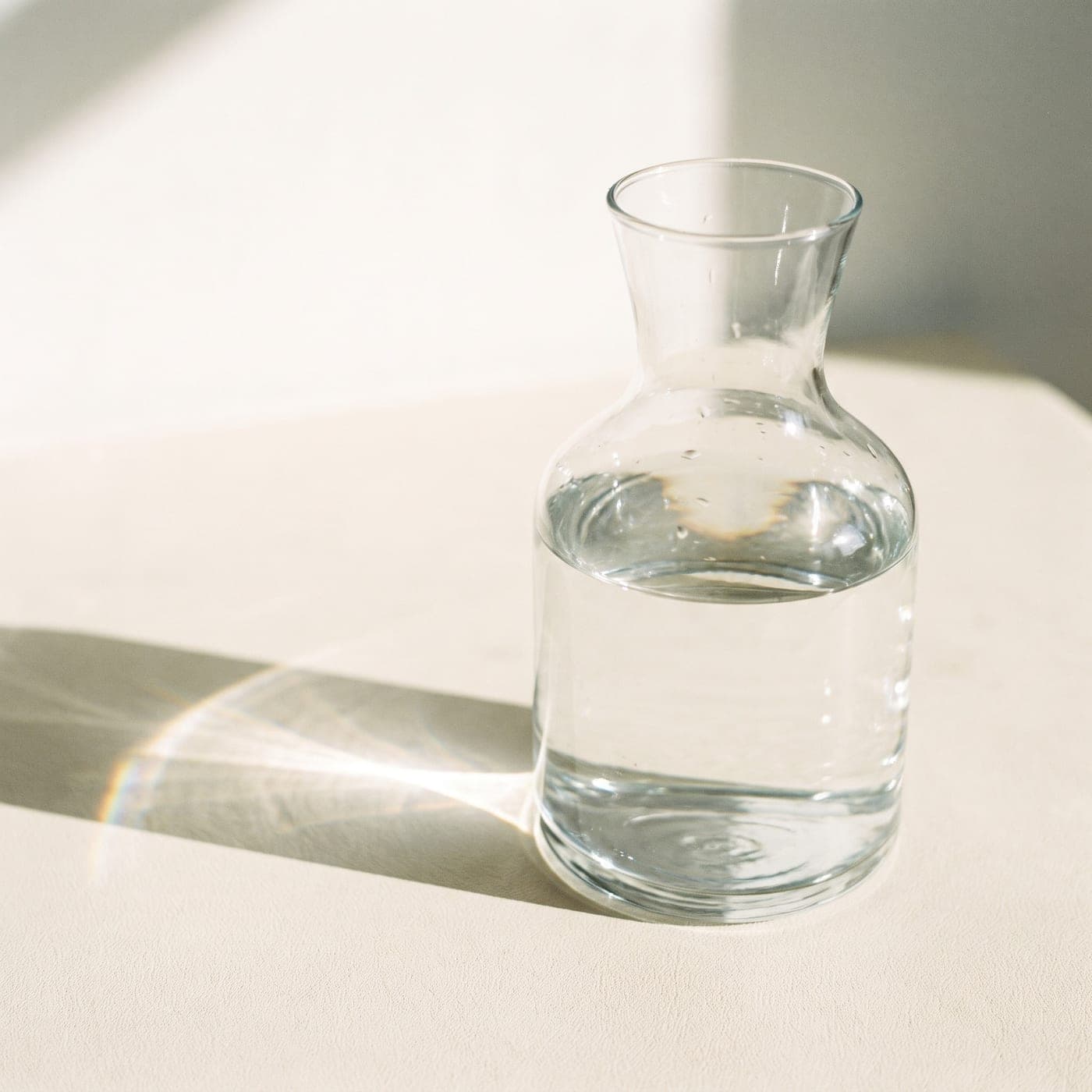 A clear glass carafe of water on a pale surface, refracting a band of daylight